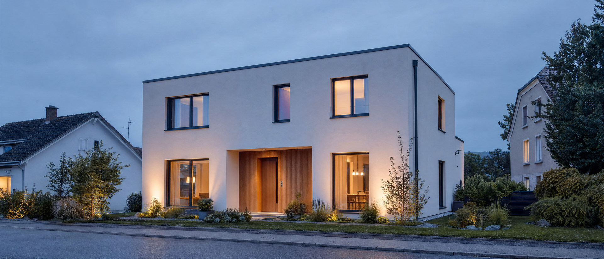 Modernes Einfamilienhaus mit heller Fassade und warm beleuchteten Fenstern in der blauen Stunde